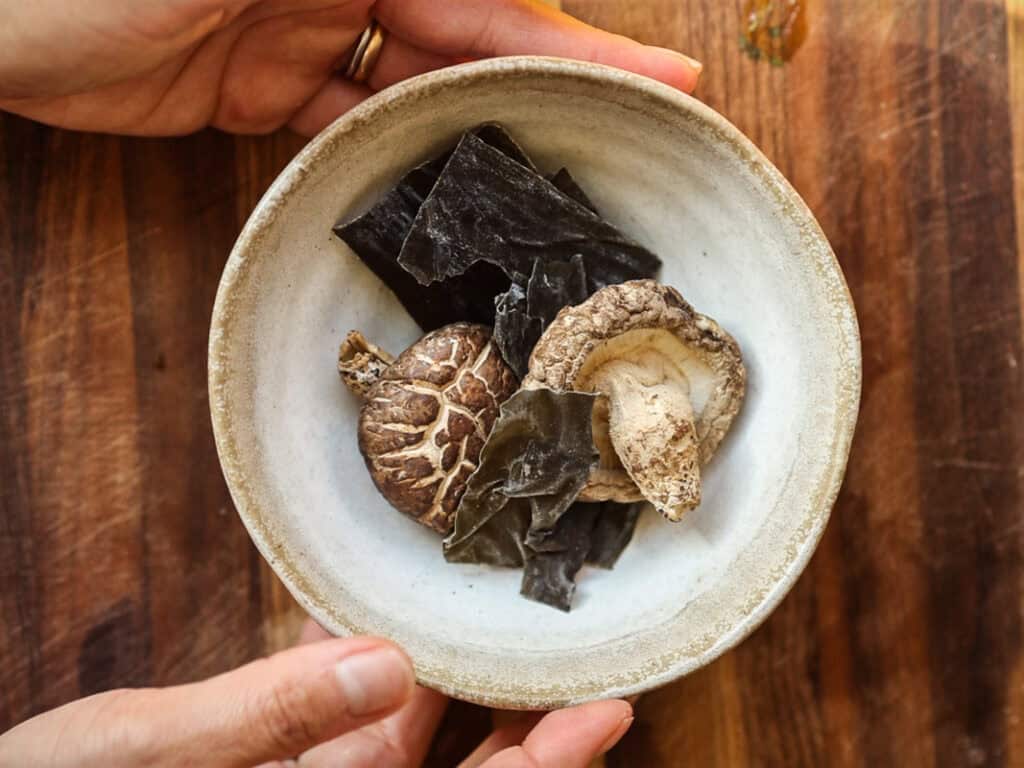 Hands holding a ceramic bowl containing dried shiitake mushrooms and seaweed, set on a wooden surface.